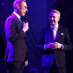 Alexander Stubb speaking into a microphone standing on a dark stage next to Pekka Haavisto, both wearing dark suits.