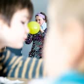 Varhaiskasvatuksen opettaja Outi Ahjos puhaltaa keltaista ilmapalloa tuima ilme kasvoillaan. Etualalla epäskarppi kuva lapsesta. 