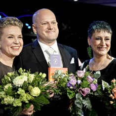 A bald man holding a book flanked by two women with short hair, all smiling and holding flowers. 