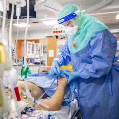 Henkilö makaa sairaalasängyssä. Suojavarusteisiin pukeutunut hoitaja asettaa potilaalle paitaa päälle.