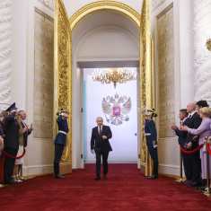 Vladimir Putin astuu saliin suurista kultaisista ovista. Hänellä on musta puku ja punainen kravatti. Oven molemmin puolin seisovat asennossa sotilaat juhlaunivormuissa ja tekevät kunniaa. Molemmin puolin käytävää seisoo taputtavaa juhlayleisöä punaisen nauhan takana.