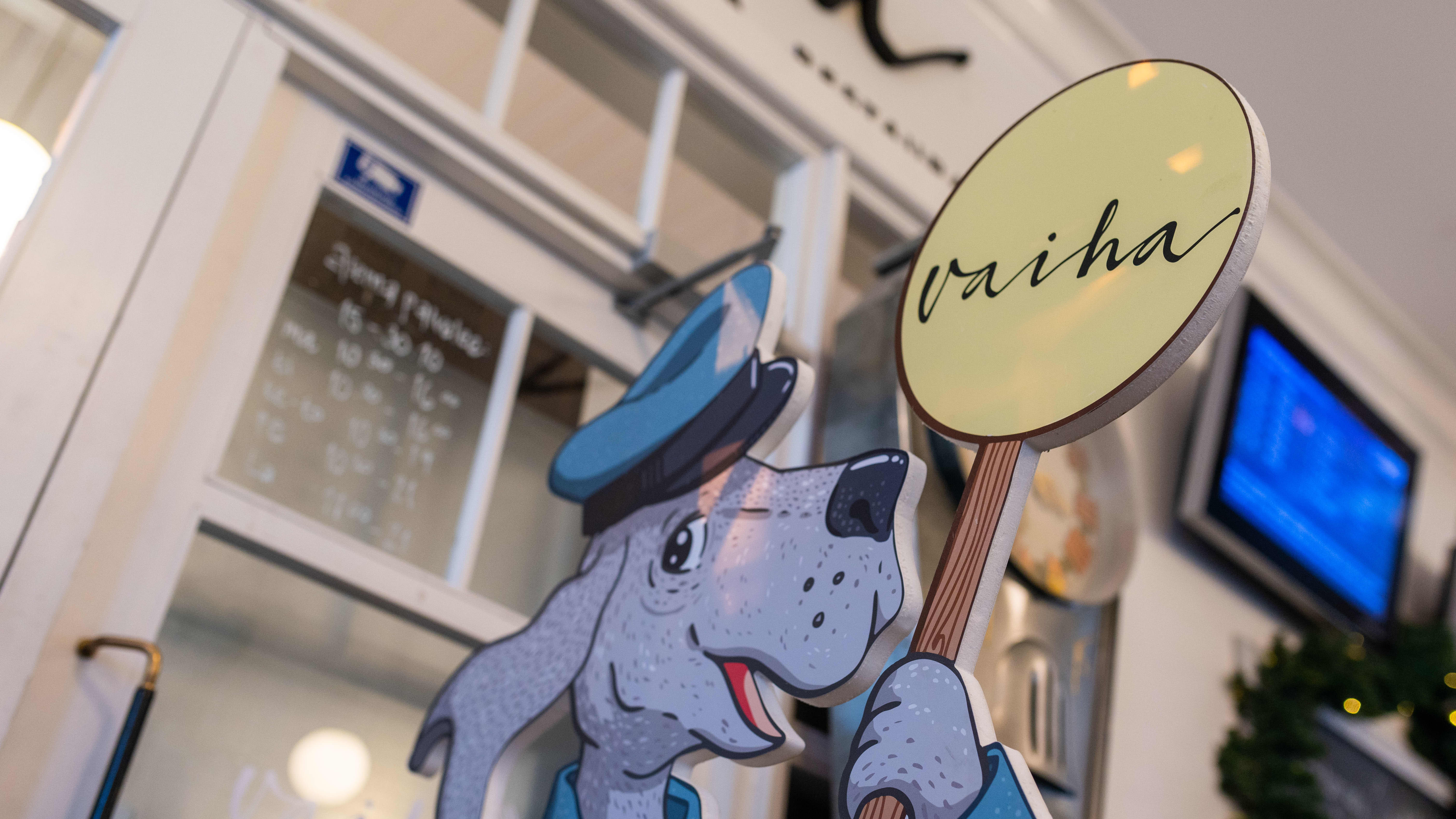 Mikkelin rautatieaseman sisätiloissa ravintola vaihan mainoskyltti. Mainoskyltissä ravintolaa mainostaa konduktooriksi pukeutunut piirretty koirahahmo.