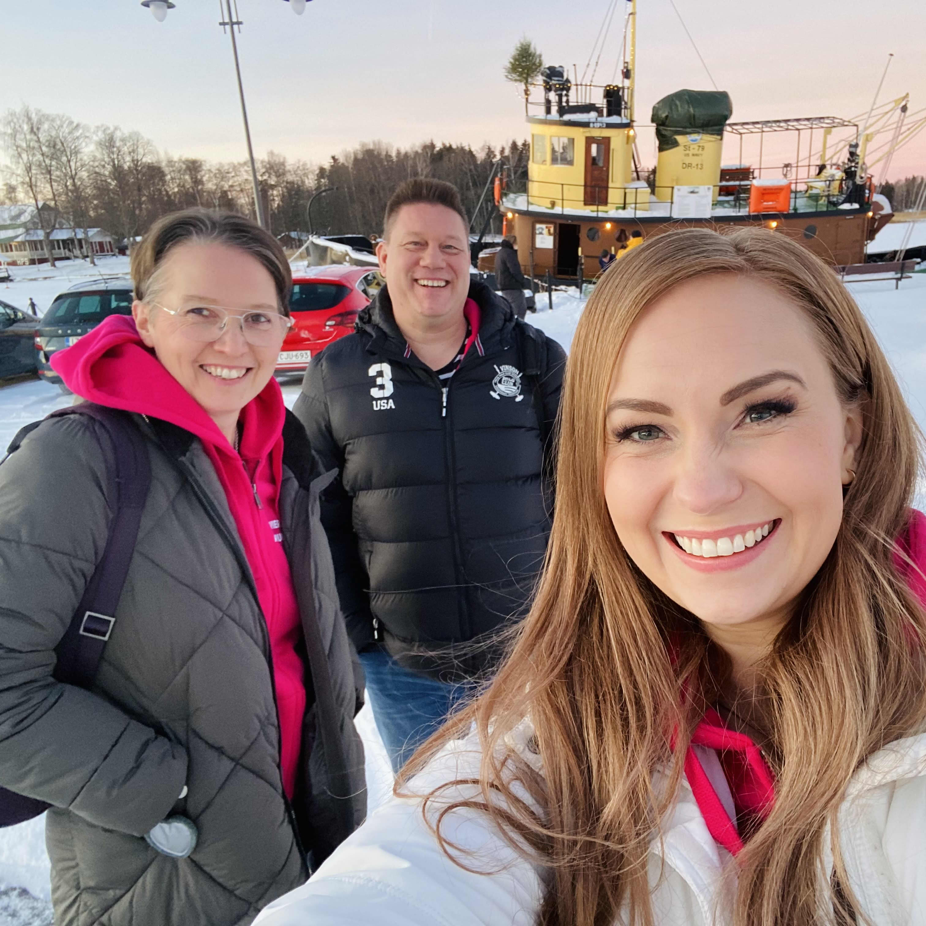 Kolme hymyilevää keski-ikäistä henkilöä seisoo lumisessa satamassa, yksi heistä kuvaa selfienä koko ryhmää.