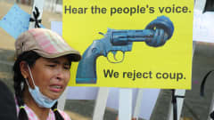 En ung kvinna med skärmmössa står framför ett gult plakat med en revolver som fått knut på pipan. Plakatet har texten "Hear the people's voice. We reject coup".