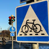 Polkupyöräilijä odottaa valojen vaihtumista Turun Aninkaistensillan kupeessa. Liikennemerkki B7, väistämisvelvollisuus pyöräilijän tienylityspaikassa. Neliönmallisessa sinipohjaisessa merkissa on valkoisen kolmion sisällä polkupyörän kuva sekä katkoviivoitus osoittamassa pyörätien jatketta.