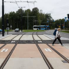 Henkilö ylittää raiteita ylityspaikalla. Taustalla autoliikennettä. Kiskot jatkuvat kohti horisonttia.