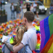Kaksi henkilöä nojaa toisiinsa Pride-lippu olalla, taustalle on laskettu kukkia.