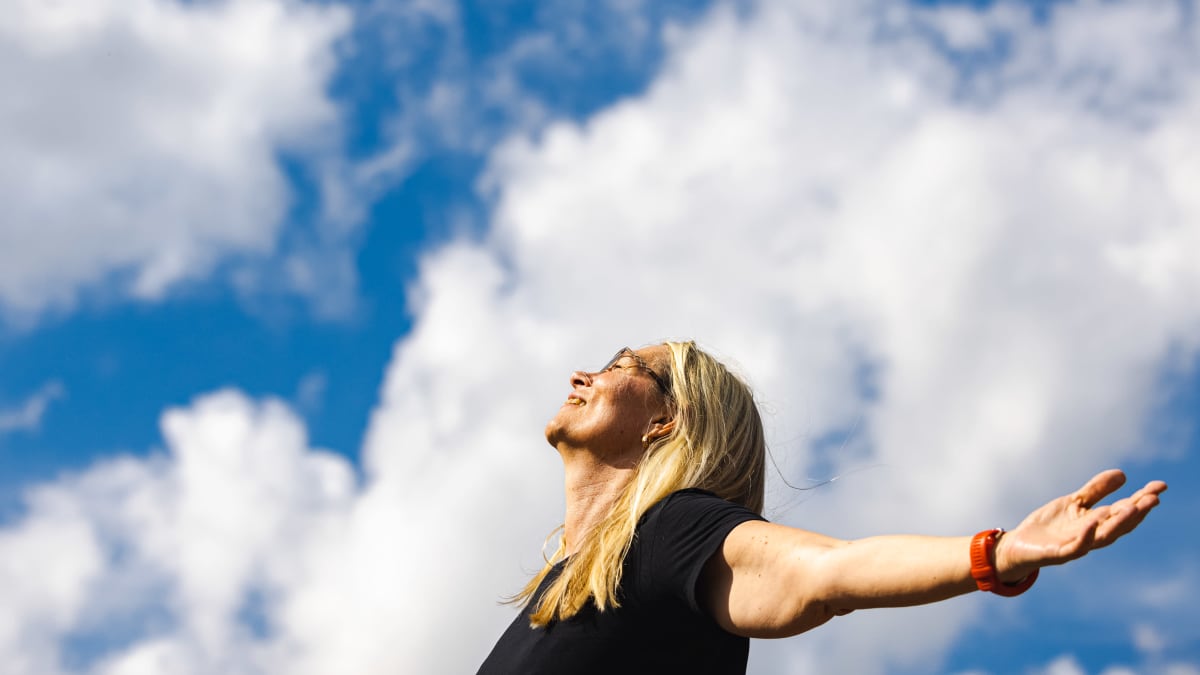 Kirsi Marie Liimatainen seisoo kädet levällään nauraen kohti taivasta katsoen taivas pilvineen taustalla.