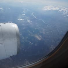 Kuva lentokoneen ikkunasta ulos; alhaalla häämöttää maisema ja asutusta.