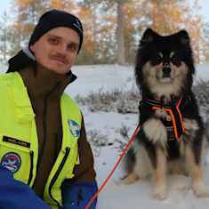 Nuori mies lumisessa metsässä keltainen huomioliivi päällä ja hänen vieressään istuu musta-vaalea koira.