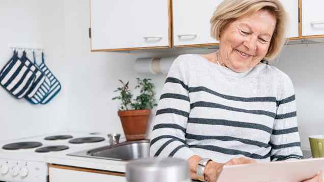 Hymyilevä nainen tutkii tablettitietokoneen ruutua keittiössä. Kuvituskuva.