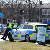 Kuvan etualalla kaksi poliisia nojaa poliisiautoon. Poliisiauton lähellä on kaupan mainoskyltti, jossa lukee testin paras.