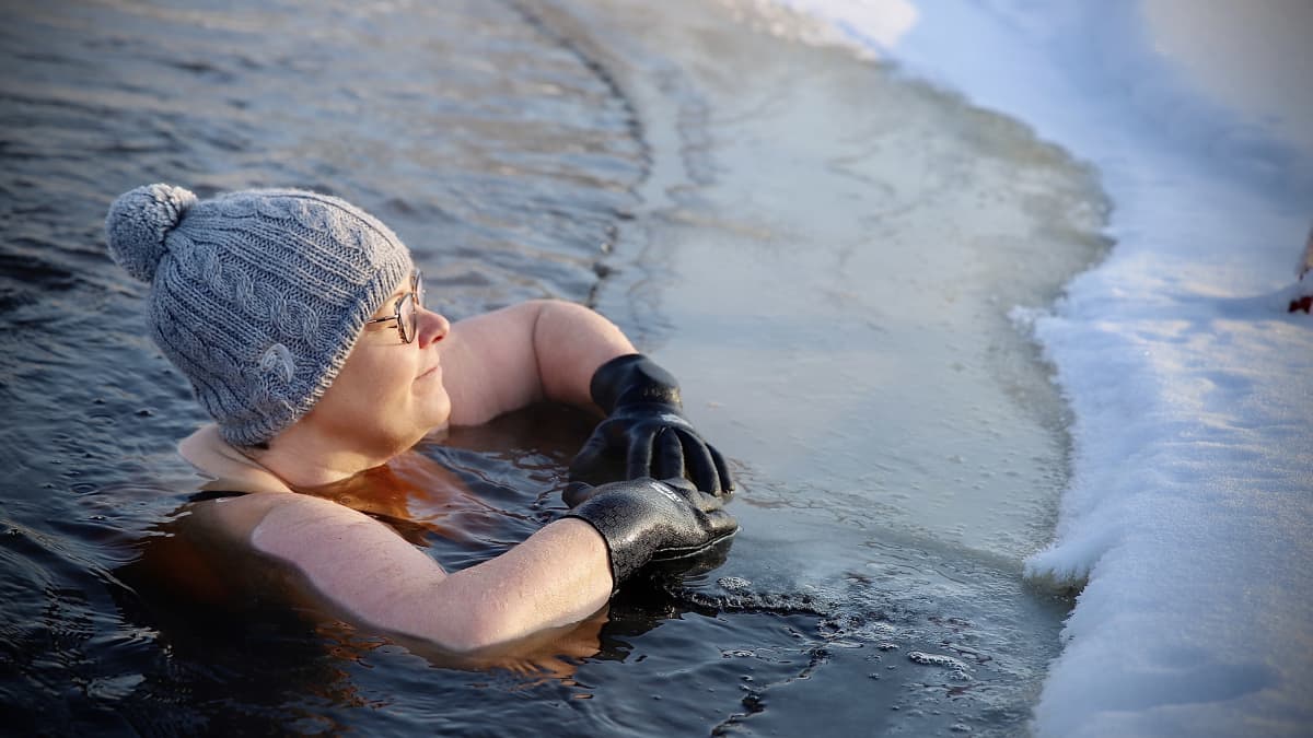 Satu-Maarit Alho pipo päässä ja hanskat kädessä avannossa nojailemassa jäätä vasten. 