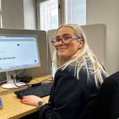 Nuori nainen istuu työpöytänsä ääressä tietokoneellaan ja katsoo kameraan. 