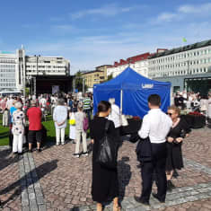 Kesävaatteisiin pukeutuneita ihmisiä torilla, jonka on päällystetty punertavin mukulakivin. Ihmiset katsovat kohti kauempana olevaa esiintymislavaa. Taustalla on kerrostaloja. 