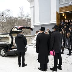 Hautajaisvieraita ja ruumisauto kirkon edustalla.