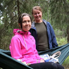 Sari Kalenius ja Tiina Martikainen hymyilevät riippukeinussa metsässä.