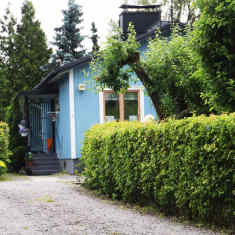 En grusväg leder fram till ett blått hus skymtar bakom grönskande träd och buskar.