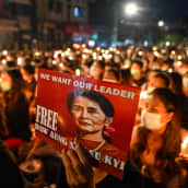 Mielenosoittaja pitää kuvaa syrjäytetystä johtajasta Aung San Suu Kyistä. Kyltin teksti vaatii vapautta Kyille. Taustalla mielenosoittajat pitelevät kynttilöitä. 