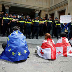 Demonstranter insvepta i EU:s och Georgiens flaggor sitter utanför en byggnad vars ingång bevakas av ett stort antal poliser.