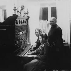 Puhelinkeskus 1920-luvulla. Kaksi puhelinoperaattoria, joiden takana seisoo nainen. Keskuksen toisella puolella johtokeskuksen luona seisoo vielä yksi mies.