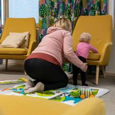 Alle vuoden ikäinen lapsi seisoo lattialla nojatuolin istuinta vasten. Hänen vieressään istuu lapsen äiti polvillaan ja pitää hellästi kiinni.