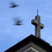 Kirkon risti ja ohi lentäviä lintuja sinistä taivasta vasten. 
