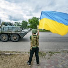 Poika heiluttaa Ukrainan lippua tienvarressa kypärä päässään panssariajoneuvon ajaessa ohi.