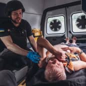Ensihoitaja pitelee haavoittunutta siviiliä ambulanssin kyydissä. Ensihoitajalla on kumihanskat, luotiliivi ja kypärä. T-paidan hihassa on Ukrainan kelta-sininen lippu.