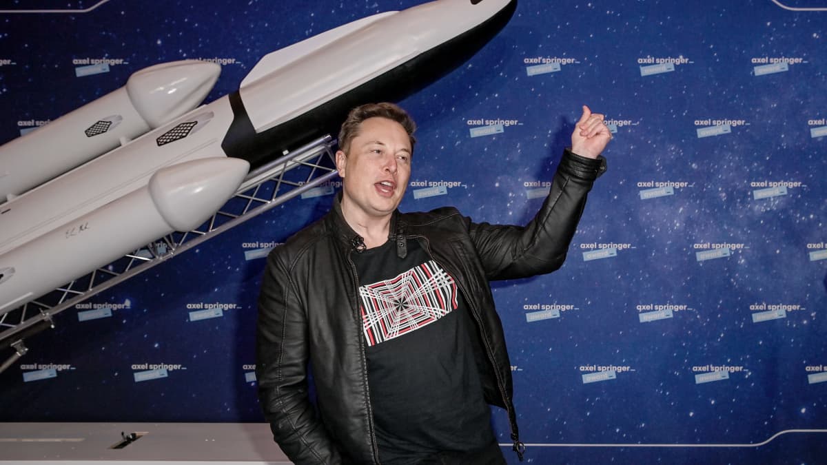 Elon Musk heiluttaa kättään, taustalla pienoismalli avaruusraketista.