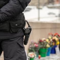 Poliisi seisoo etualalla. Kuvassa näkyy hänen vyöllään oleva pistooli. Kauempana näkyy Nemtsovin muistopaikka, jonne ihmiset tuovat kukkia.