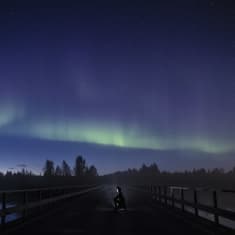 Revontulia taivaalla, mies polvillaan sillalla valoa kasvoilla.