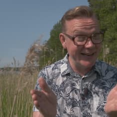 Puoli seitsemän -ohjelman toimittaja Marko Pulkkinen seisoo kesäisessä rantaheinikossa ja virnistää iloisesta kameralle.
