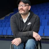 Näyttelijä Pekka Heikkinen istuu pyörätuolissa sinisen teatterikatsomon edessä.