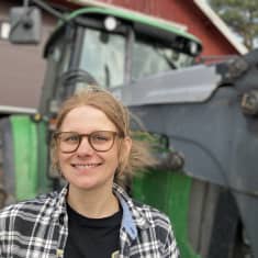 Nainen seisoo traktorin edessä ja hymyilee suoraan kameralle.