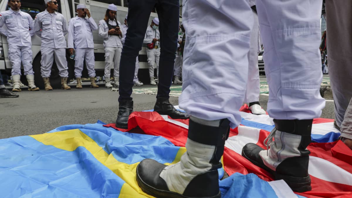 En person i vit dräkt står med den ena foten på Danmarks flagga, och den andra på Sveriges flagga.