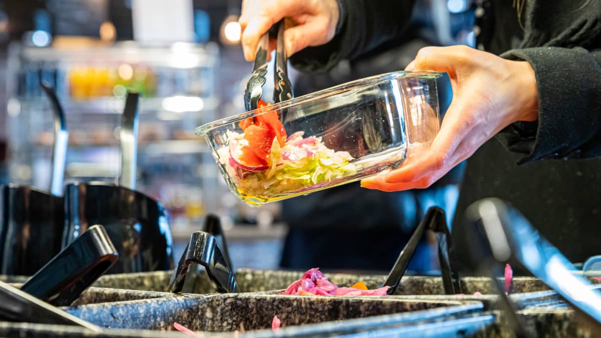 Heli kauhomassa astiaansa salattia opiskelijaravintolan seisovasta lounaspöydästä.