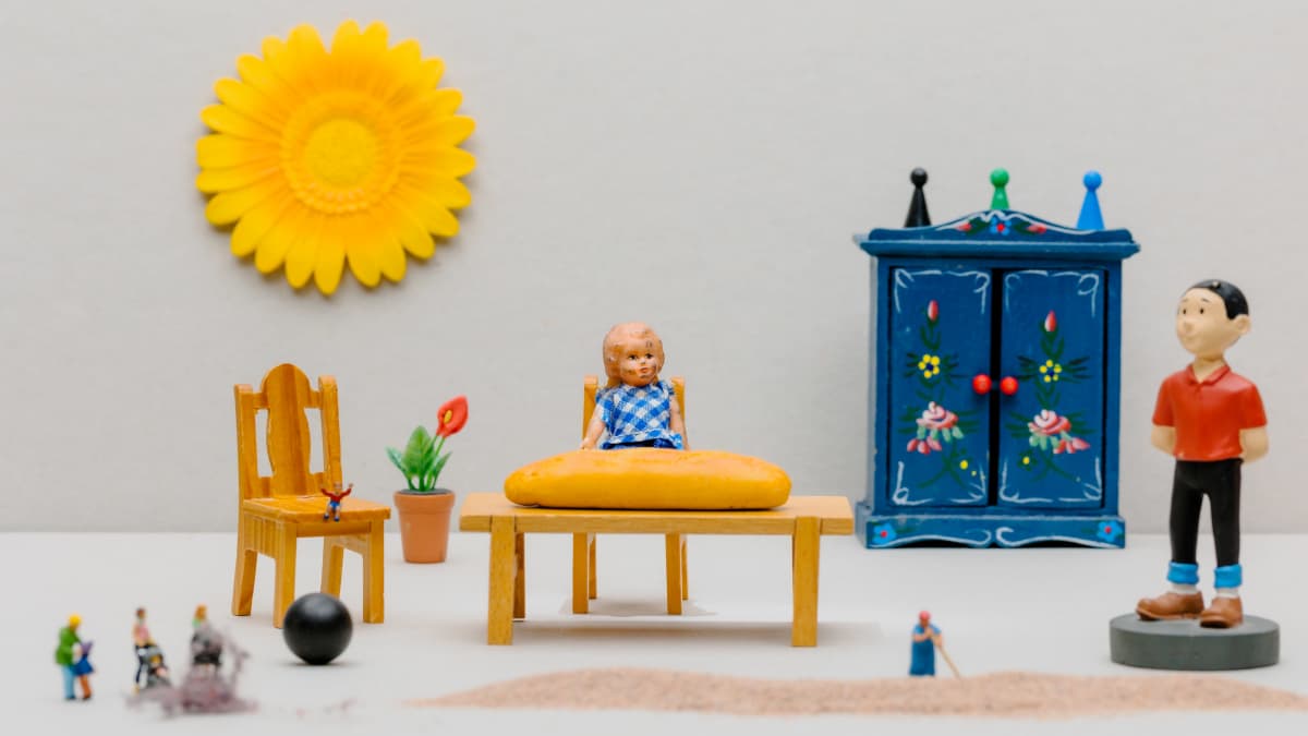 Miniatyyrimaisemassa pieni lapsi istuu pöydän ääressä, pieniä hahmoja seisoo pölyn keskellä, toinen hahmo siivoaa suurta hiekkakasaa, miestä esittävä hahmo katsoo kaikkea sivusta.
