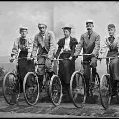 Vanhassa mustavalkoisessa valokuvassa vuosisadan alun nuori naisia ja miehiä polkupyörillä rivissä.  