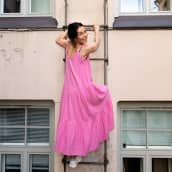 Nainen vaaleanpunaisessa kesämekossa kiipeää tikapuita ylös ja hymyilee. Takana kerrostalon seinä ja ikkunoita. 