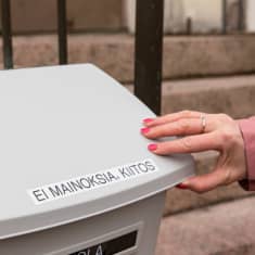 Lähikuva vaalean muovisen postilaatikon kannesta jossa olevassa tarrassa lukee ei mainoksia, kiitos. Kuvan oikeassa laidassa naisen käsi pitää kiinni kannesta.