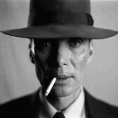 Svartvit närbild på J. Robert Oppenheimer som är klädd i svart kostym, vit skjorta och hatt, han har en cigarett i mungipan och ser rakt in i kameran.