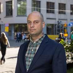 A bald man wearing a plaid shirt and blue blazer stands by a sunny city street with pedestrians behind him.