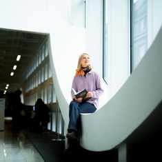 Nuori nainen pitelee kirjaa ja istuu ison ikkunan ääressä kirjastossa.