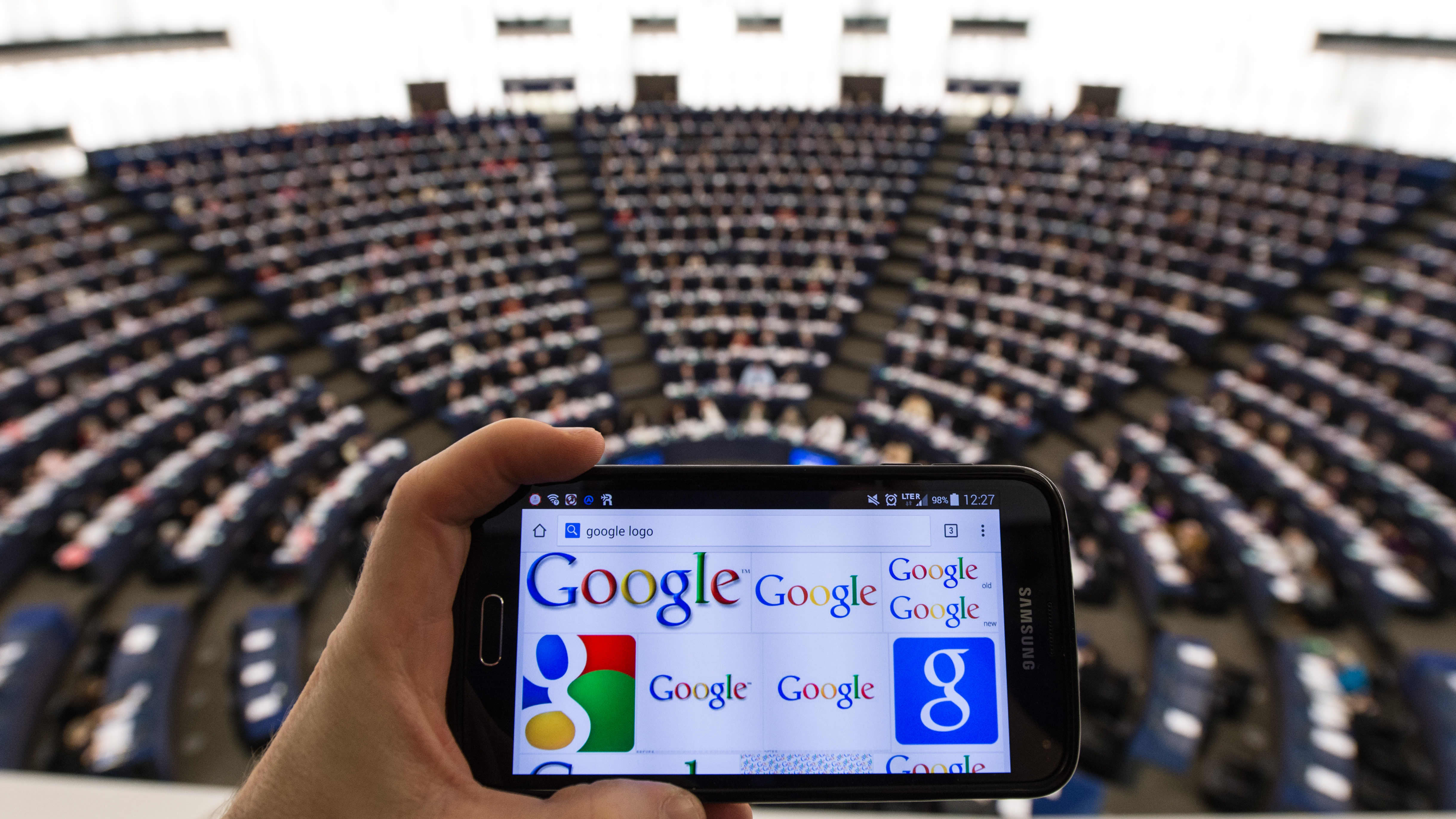 Euroopan parlamentin sali, jonka edessä Googlen-logo kännykän näytöllä.
