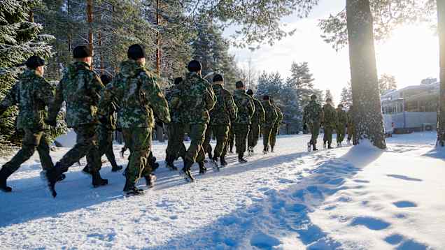 A large group of soldiers in camouflage uniforms marching in formation in the snow, with trees and a sunny sky in the background.
