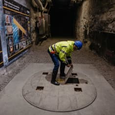 Man wearing bright yellow jacket and blue helmet, lifting a metal door on the floor of a long, rocky tunnel.