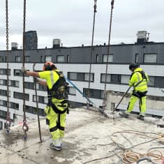 Kaksi henkilöä seisoo rakennustyömaalla paljaalla betonilattialla. Taustalla näkyy kerrostaloja.