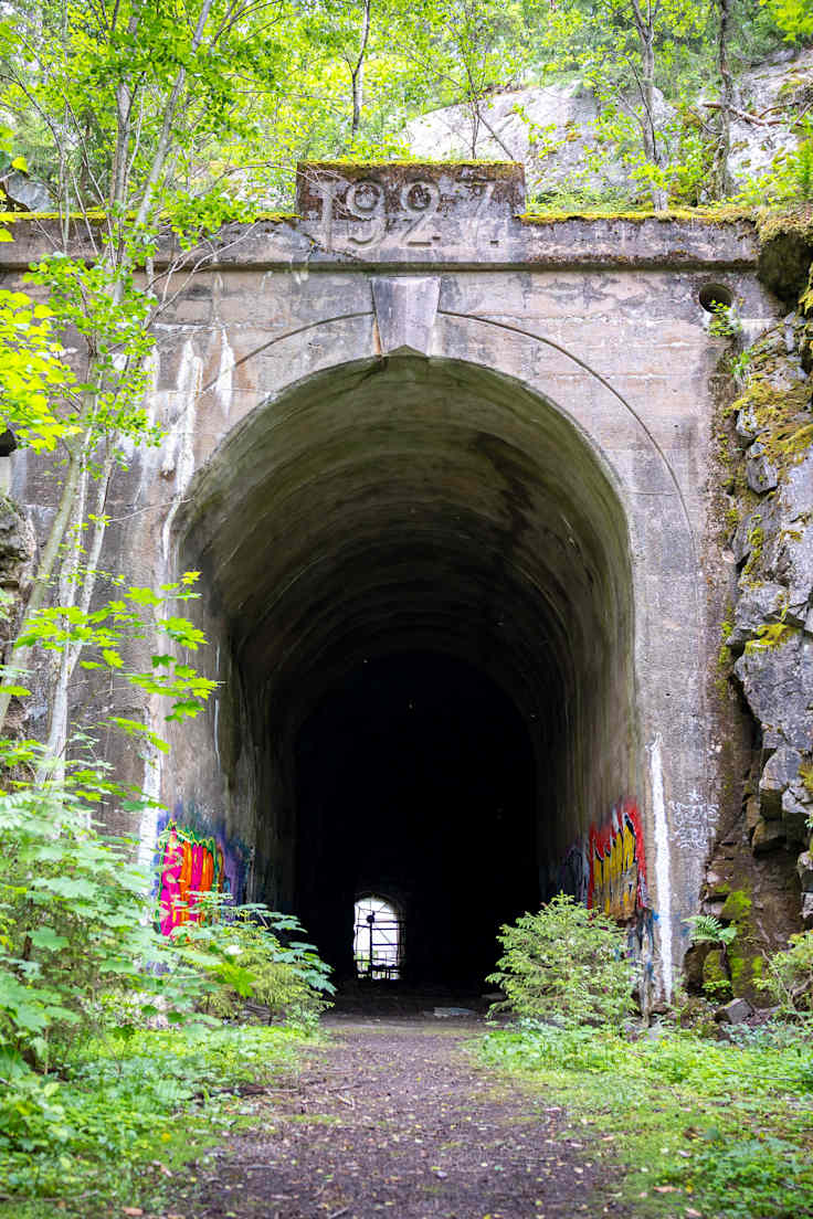 Hylätyn rautatietunnelin korkea sisäänkäynti. Ympärillä on vehreää kasvillisuutta. Tunnelin päällä on kaiverrettu teksti 1927.