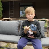 Poika istuu puutarhatuolilla ja pitelee käsissään kyniä ja penaalia. Kuvan vasemmassa reunassa on kukkia ruukussa. 